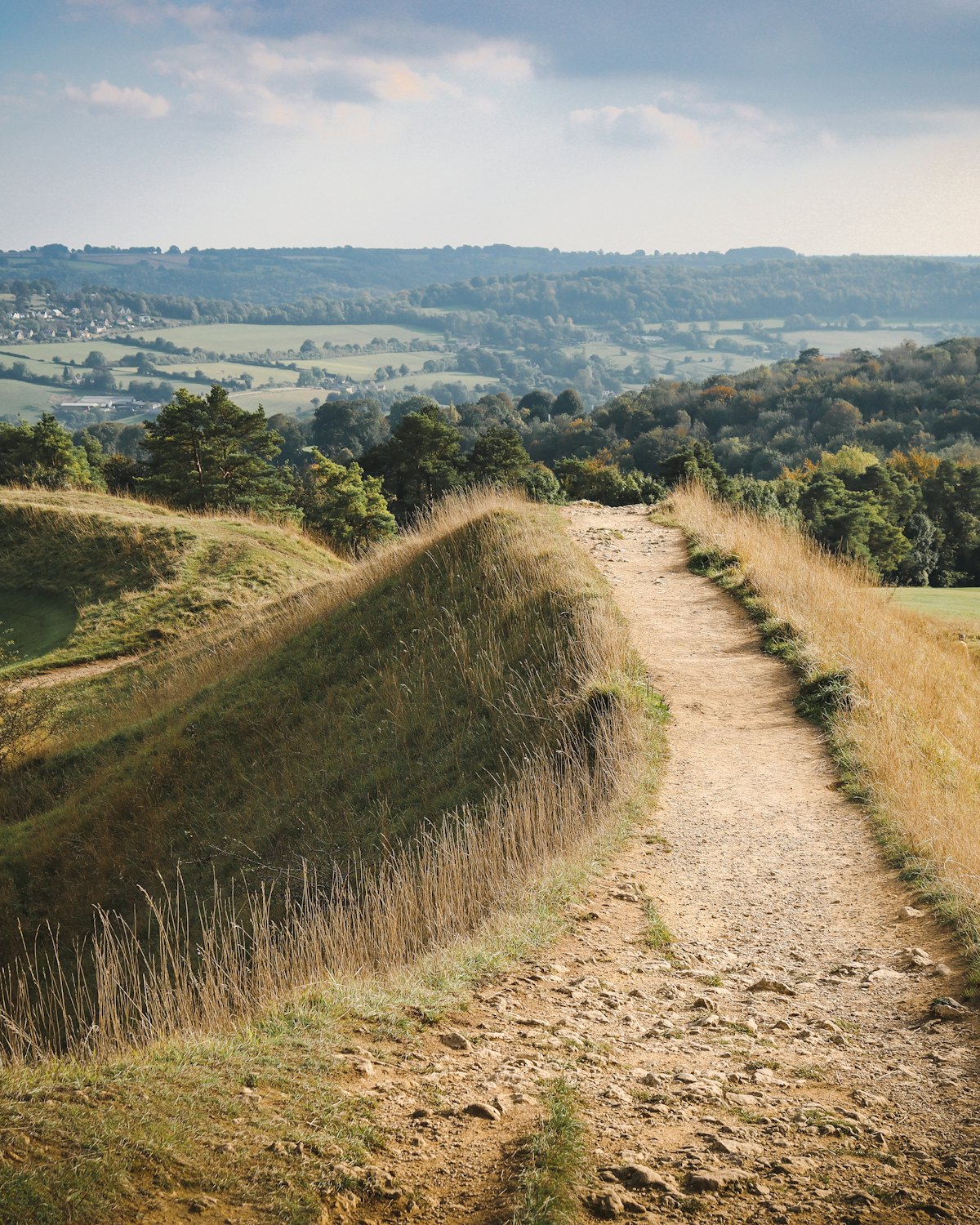 A footpath running along a grassy Cotswold ridge, with wooded hills in the distance